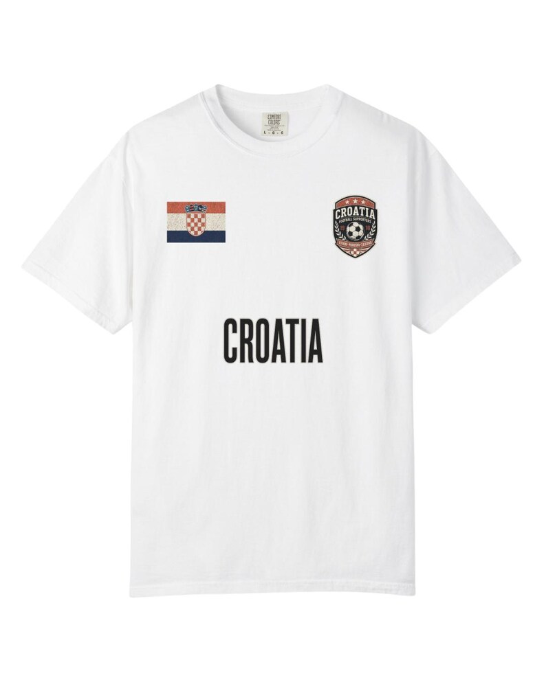 May include: White t-shirt featuring the word "CROATIA" in large black letters. The shirt has a Croatian flag patch on the left chest and a soccer-themed emblem on the right chest. The shirt is a casual wear item.