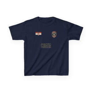 May include: Navy blue t-shirt featuring the word "CROATIA" in a gold font. The shirt has a Croatian flag patch and a soccer crest. The shirt is a solid color and has short sleeves.
