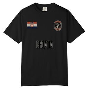 May include: Black t-shirt featuring the word "CROATIA" in white block letters. The shirt has a Croatian flag patch on the left sleeve and a "CROATIA" crest with a soccer ball on the right chest.