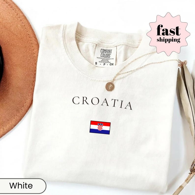 Peut inclure: T-shirt blanc avec le mot "CROATIA" imprim&eacute; en noir et un petit drapeau croate. Le t-shirt a un col rond et des manches courtes. Un collier dor&eacute; avec un pendentif est drap&eacute; sur le t-shirt.