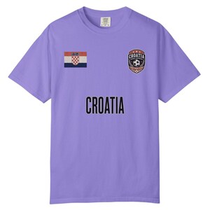May include: A lavender t-shirt featuring the word "CROATIA" in bold black letters. The shirt has a Croatian flag patch and a crest with a soccer ball and the word "CROATIA".