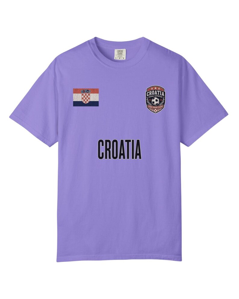 May include: A lavender t-shirt featuring the word "CROATIA" in bold black letters. The shirt has a Croatian flag patch and a crest with a soccer ball and the word "CROATIA".