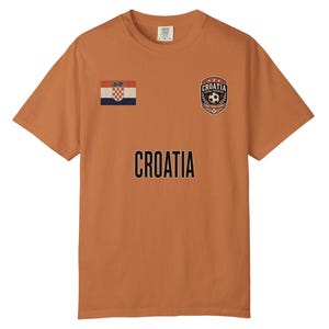 May include: An orange t-shirt featuring the word "CROATIA" in black block letters. The shirt has a Croatian flag patch and a soccer ball emblem. The shirt is short-sleeved and made of a soft material.