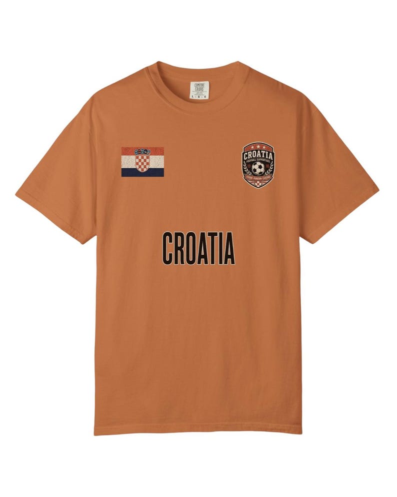 May include: An orange t-shirt featuring the word "CROATIA" in black block letters. The shirt has a Croatian flag patch and a soccer ball emblem. The shirt is short-sleeved and made of a soft material.