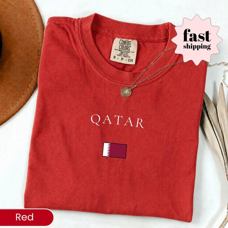 Może przedstawiać: Czerwona koszulka z napisem "QATAR" w kolorze białym i flagą Kataru. Koszulka ma okrągły dekolt i kr&oacute;tkie rękawy. Widoczny jest złoty naszyjnik z wisiorkiem.