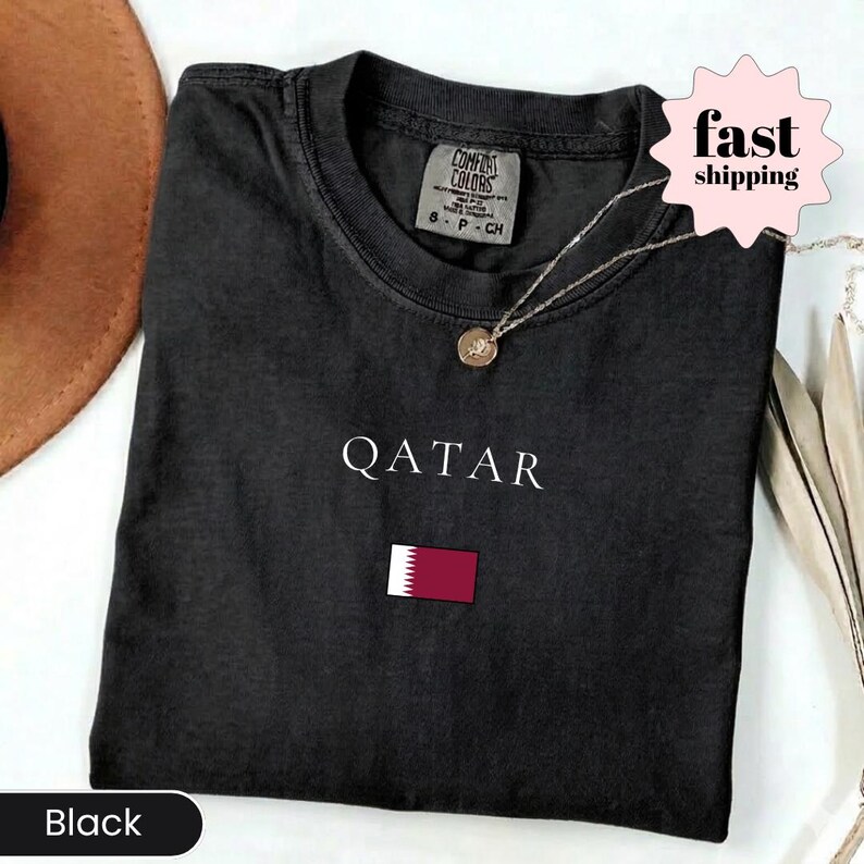Może przedstawiać: Czarny t-shirt z napisem "QATAR" w białym kolorze i małą flagą Kataru. Widoczny jest złoty naszyjnik. Koszulka jest złożona. Etykieta głosi "Comfort Colors".