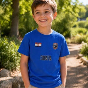 May include: A blue t-shirt with the word "CROATIA" printed across the chest. The shirt features the Croatian flag and a soccer ball emblem. The boy is wearing khaki pants and is smiling.