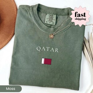 Może przedstawiać: Zielona koszulka w kolorze mchu z napisem "QATAR" w kolorze białym i małą flagą Kataru. Koszulka ma okrągły dekolt i kr&oacute;tkie rękawy. Złoty naszyjnik z wisiorkiem znajduje się na koszulce.