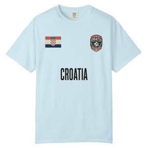 May include: Light blue t-shirt featuring the word "CROATIA" in bold black letters. The shirt has a Croatian flag patch and a soccer-themed emblem. A comfortable, casual top for fans of Croatian soccer.