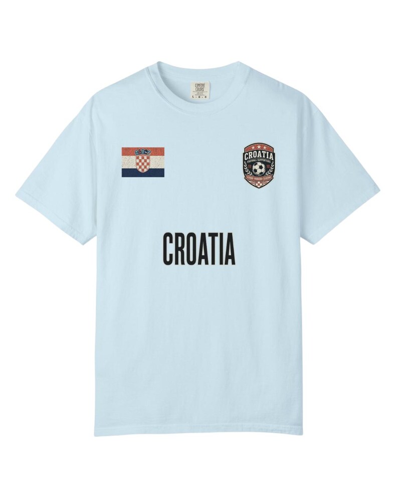 May include: Light blue t-shirt featuring the word "CROATIA" in bold black letters. The shirt has a Croatian flag patch and a soccer-themed emblem. A comfortable, casual top for fans of Croatian soccer.