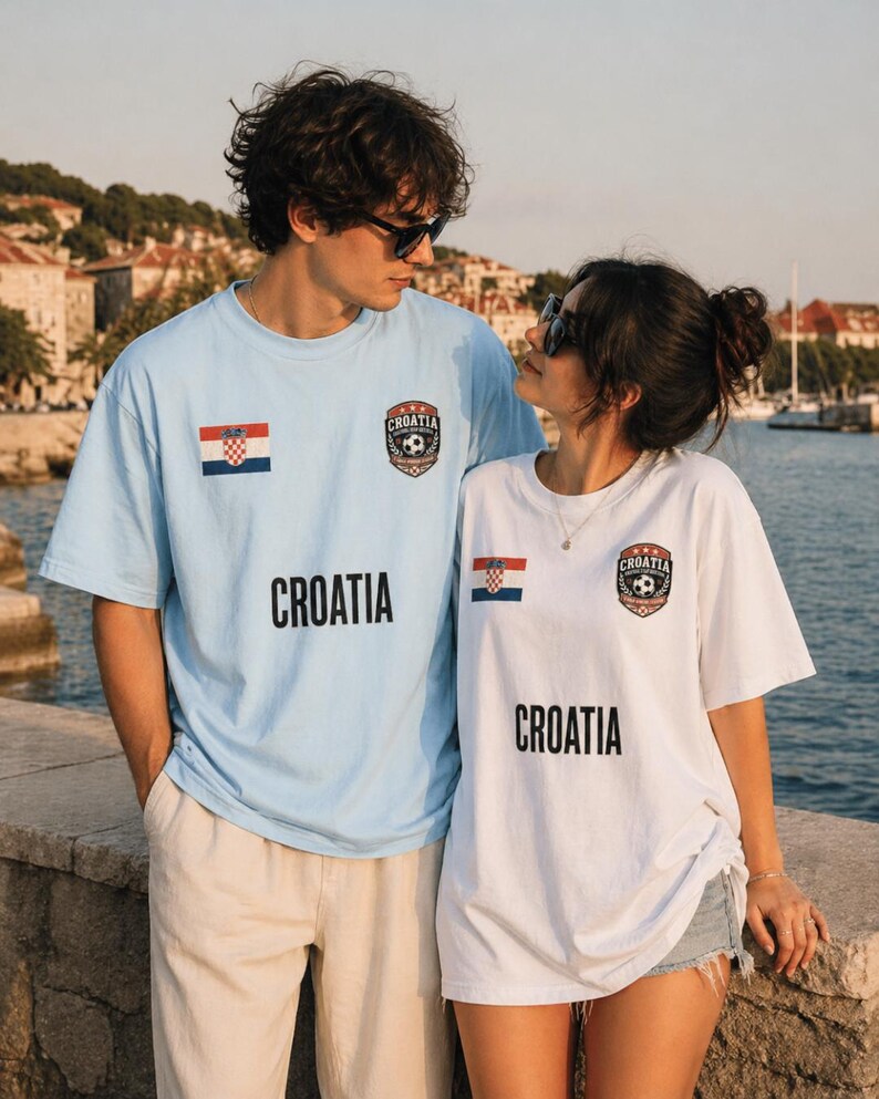 May include: Light blue and white Croatia t-shirts. Each shirt features the Croatian flag and the word "CROATIA" in black block letters. A soccer ball emblem is also present. The shirts are short-sleeved and worn by two people.