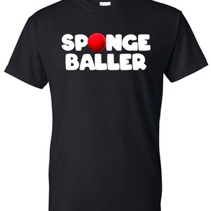 May include: Black t-shirt with the words "SPONGE BALLER" in large, white, uppercase letters. A red sphere is positioned above the word "BALLER". The shirt is a crew neck style.