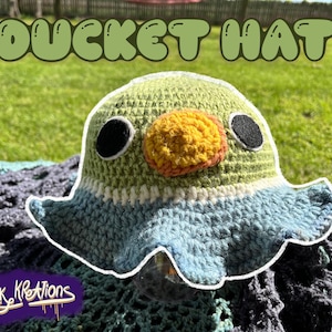 May include: A handmade crochet bucket hat in the shape of a duck. The hat features a green and blue color scheme with a yellow beak and black eyes. The text "DUCKET HAT" is displayed above the hat.