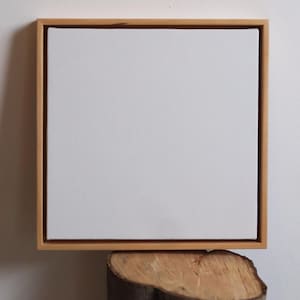Puede incluir: Un lienzo blanco cuadrado en blanco estirado sobre un marco de madera. El marco tiene un acabado de madera natural. El lienzo descansa sobre una sección de un tronco de árbol, con una pared blanca en el fondo. La obra de arte está lista para pintar.