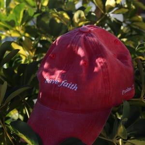 以下が含まれることがあります： 「have faith」と「Psalm 21」の刺繍が入った赤い野球帽。柔らかい素材でできており、つばがカーブしています。背景には緑と黄色の葉があります。