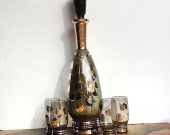 May include: A set of four clear glass shot glasses with gold leaf accents and a matching green glass decanter with a gold leaf design and a black stopper.