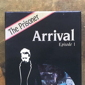 May include: A black VHS tape with a white and red label that reads "The Prisoner Arrival Episode 1 starring Patrick McGoohan". The image on the tape is a black and white silhouette of a man in a suit standing in front of a circular image of a group of people walking under umbrellas.