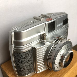 Może przedstawiać: Srebrny aparat fotograficzny w stylu vintage z teksturowanym uchwytem i obiektywem z ustawieniami odległości. Aparat posiada mocowanie lampy błyskowej i pokrętło z numerami. Aparat stoi na drewnianej powierzchni.