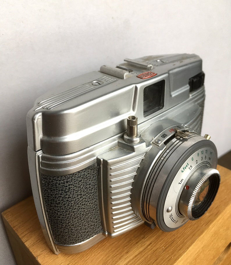 Może przedstawiać: Srebrny aparat fotograficzny w stylu vintage z teksturowanym uchwytem i obiektywem z ustawieniami odległości. Aparat posiada mocowanie lampy błyskowej i pokrętło z numerami. Aparat stoi na drewnianej powierzchni.
