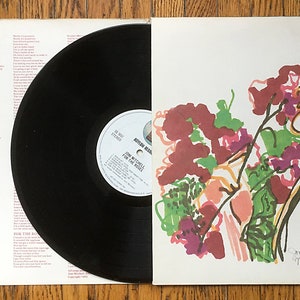 May include: A white vinyl record album cover with a colorful abstract painting of a woman smelling flowers. The album title is "For the Roses" by Joni Mitchell.