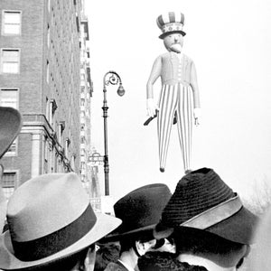 May include: A black and white photo of a crowd of people looking up at a large inflatable Uncle Sam figure. The figure is wearing a red, white, and blue striped suit and a hat with stars. The photo was taken in a city street with tall buildings in the background.