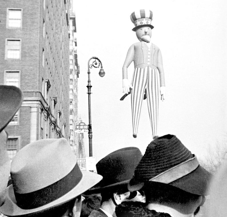 May include: A black and white photo of a crowd of people looking up at a large inflatable Uncle Sam figure. The figure is wearing a red, white, and blue striped suit and a hat with stars. The photo was taken in a city street with tall buildings in the background.