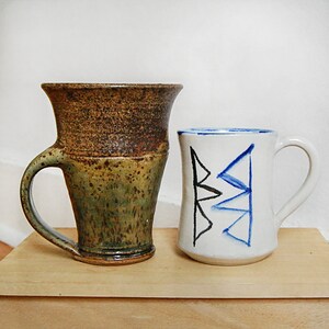May include: Two ceramic mugs on a wooden surface. The mug on the left is brown and green with a wide handle. The mug on the right is white with blue and black geometric designs and a curved handle. Both mugs are handmade.