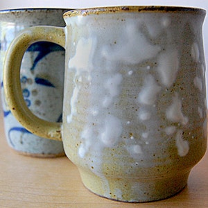 Puede incluir: Dos tazas de cer&aacute;mica hechas a mano. La taza delantera es de color beige claro con manchas blancas y un asa amarilla. La segunda taza es azul claro con un dise&ntilde;o floral azul. Ambas tazas tienen un aspecto r&uacute;stico y artesanal.