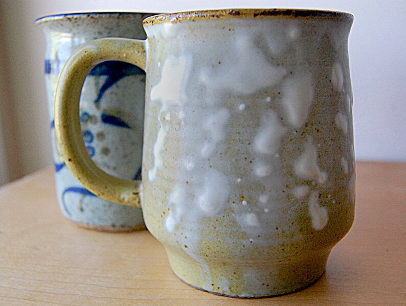 Puede incluir: Dos tazas de cer&aacute;mica hechas a mano. La taza delantera es de color beige claro con manchas blancas y un asa amarilla. La segunda taza es azul claro con un dise&ntilde;o floral azul. Ambas tazas tienen un aspecto r&uacute;stico y artesanal.