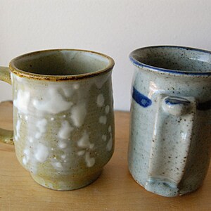 Puede incluir: Dos tazas de cer&aacute;mica hechas a mano. La taza de la izquierda es de color verde claro con detalles blancos y un asa redondeada. La taza de la derecha es azul claro con una franja azul y un asa cuadrada. Ambas tazas est&aacute;n sobre una superficie de madera.