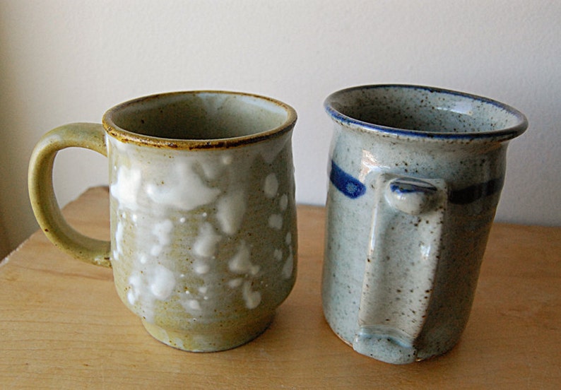 Puede incluir: Dos tazas de cer&aacute;mica hechas a mano. La taza de la izquierda es de color verde claro con detalles blancos y un asa redondeada. La taza de la derecha es azul claro con una franja azul y un asa cuadrada. Ambas tazas est&aacute;n sobre una superficie de madera.