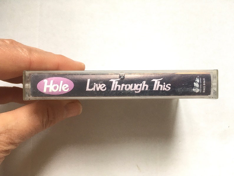 May include: A clear plastic cassette tape case with a black background and white text. The text reads "Hole Live Through This".