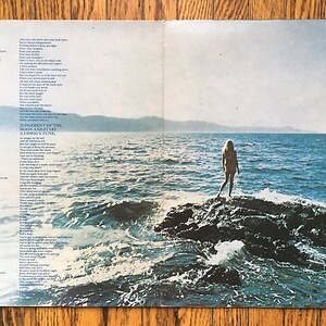 May include: A vintage record album cover featuring a woman standing on a rock in the ocean. The album cover is a light blue color with a bright blue sky and a light blue ocean. The woman is wearing a white bikini and has long blonde hair. The album cover has lyrics printed on the left and right sides.