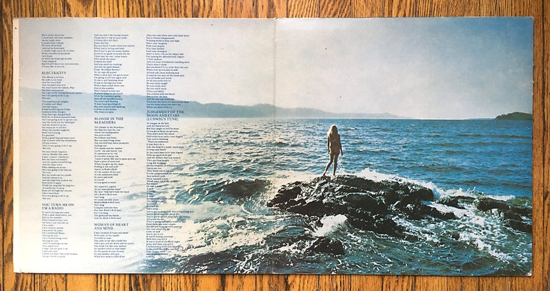 May include: A vintage record album cover featuring a woman standing on a rock in the ocean. The album cover is a light blue color with a bright blue sky and a light blue ocean. The woman is wearing a white bikini and has long blonde hair. The album cover has lyrics printed on the left and right sides.