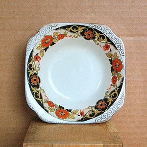 May include: A square, white ceramic bowl with a floral design. The rim features a black and gold border with orange flowers and gold accents. The bowl sits on a wooden surface.