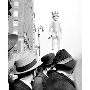 May include: A black and white photo of a crowd of people looking up at a large inflatable Uncle Sam figure in the sky. The people are wearing hats and coats.
