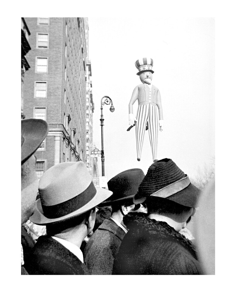 May include: A black and white photo of a crowd of people looking up at a large inflatable Uncle Sam figure in the sky. The people are wearing hats and coats.