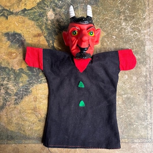 May include: A hand puppet featuring a molded red devil head with white horns, yellow eyes, and a black goatee. The body is made of black fabric with red sleeves, a red triangular collar piece, and two small green felt triangles on the front.