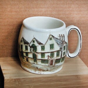 May include: A white ceramic mug with a brown handle. The mug features a hand-painted image of a building with green windows and a brown roof. The building is surrounded by a green lawn and trees.