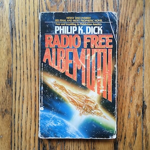 May include: A paperback book cover with a blue, black, and orange color scheme. The title is "Radio Free Albemuth" by Philip K. Dick. The cover features a spaceship flying over a planet.