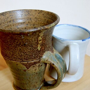 May include: Two ceramic mugs. One is a large, textured mug with a brown and green glaze and a curved handle. The other is a smaller, white mug with a blue rim and a white handle. Both mugs are on a light-colored surface.