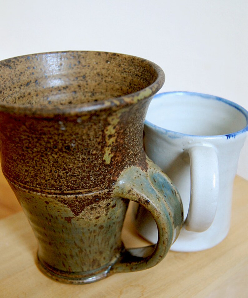 May include: Two ceramic mugs. One is a large, textured mug with a brown and green glaze and a curved handle. The other is a smaller, white mug with a blue rim and a white handle. Both mugs are on a light-colored surface.