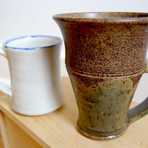 May include: Two ceramic mugs. One is white with a blue rim, the other is brown and green with a textured surface. Both mugs have handles and are on a wooden surface.
