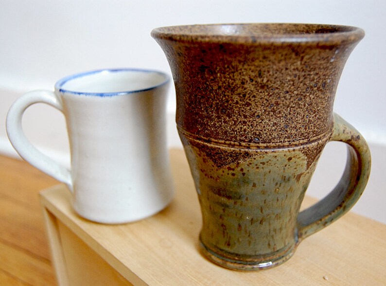 May include: Two ceramic mugs. One is white with a blue rim, the other is brown and green with a textured surface. Both mugs have handles and are on a wooden surface.
