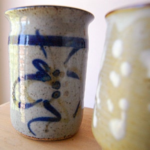 Puede incluir: Dos jarrones de cer&aacute;mica hechos a mano. El jarr&oacute;n enfocado es cil&iacute;ndrico, con un esmalte moteado gris y beige. Presenta un dise&ntilde;o abstracto azul. El segundo jarr&oacute;n est&aacute; desenfocado, con un esmalte amarillo claro y manchas blancas.