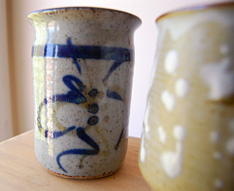 Puede incluir: Dos jarrones de cer&aacute;mica hechos a mano. El jarr&oacute;n enfocado es cil&iacute;ndrico, con un esmalte moteado gris y beige. Presenta un dise&ntilde;o abstracto azul. El segundo jarr&oacute;n est&aacute; desenfocado, con un esmalte amarillo claro y manchas blancas.