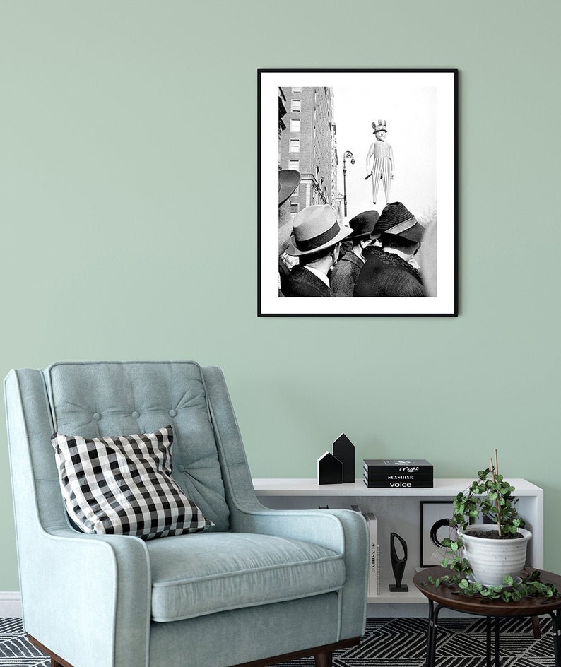 May include: Black and white photograph of three men in hats looking up at a large, striped, inflatable figure of a man wearing a hat. The figure is floating in the air above a city street.