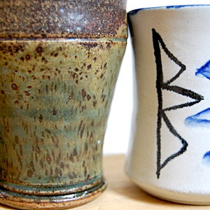 May include: Two ceramic mugs. One is a rustic, handmade mug with a green and brown speckled glaze and a handle. The other is a white mug with a black and blue geometric design. Both mugs are on a light background.