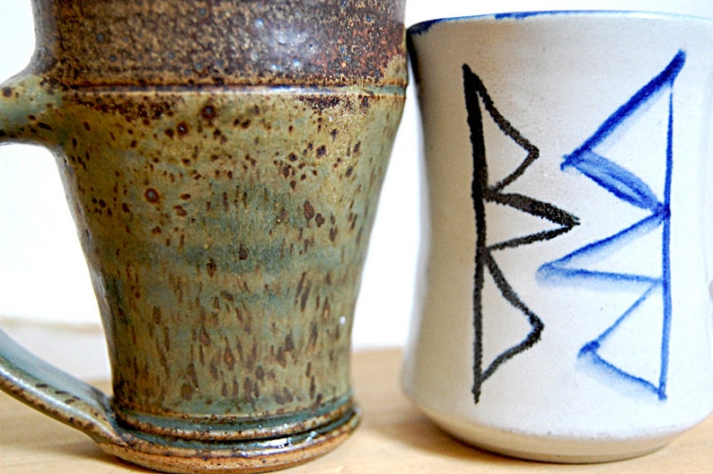May include: Two ceramic mugs. One is a rustic, handmade mug with a green and brown speckled glaze and a handle. The other is a white mug with a black and blue geometric design. Both mugs are on a light background.