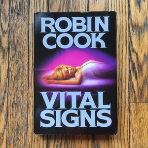 Peut inclure: Une couverture de livre de poche avec un fond noir et du texte blanc. Le titre est "Vital Signs" de Robin Cook. La couverture représente une femme allongée sur un lit avec une lueur violette autour d'elle.